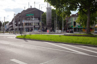 Eine Stadtkreuzung mit der BW Bank und anderen Geschäften, einem grasbewachsenen Mittelstreifen, Ampeln, geparkten Autos und einem Uhrenturm an einem teilweise bewölkten Tag.