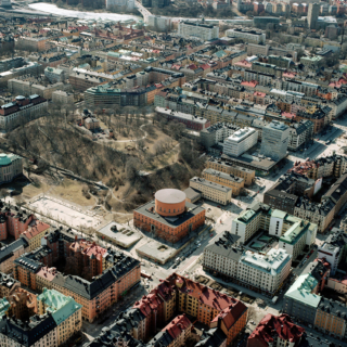 Luftaufnahme einer Stadt mit einem markanten runden Gebäude im Zentrum, umgeben von Parks, Straßen und dicht gedrängten mehrstöckigen Gebäuden.