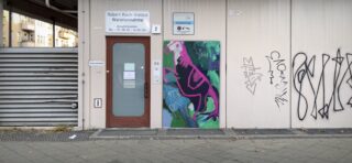 Eingang zu einem Gebäude mit Beschilderung für das Robert Koch-Institut Warenannahme, einer Tür, einem farbenfrohen rosa Dinosaurier-Wandbild und Graffiti auf einer weißen Wand.