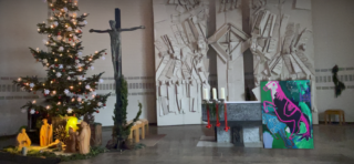 Ein Kirchenaltar ist mit einem Weihnachtsbaum, einer Weihnachtskrippe, einem Kruzifix, Kerzen und einem farbenfrohen abstrakten Gemälde eines Dinosauriers geschmückt.