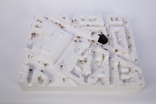 Ein weißes Architekturmodell eines Häuserblocks mit überall verstreuten kleinen Figuren und einem einzelnen schwarzen Gebäude in der Nähe der Mitte.