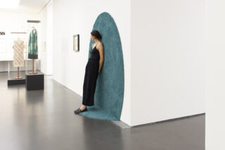 Eine Frau lehnt an einer weißen Galeriewand, die teilweise von einem blauen Strukturstoff umhüllt ist, der sich vom Boden bis zur Wand erstreckt. Im Hintergrund sind Kunstwerke ausgestellt.