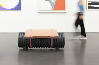 Eine Person geht in einer Kunstgalerie mit abstrakten Gemälden an der Wand an einer modernen Bank aus gerippten schwarzen Zylindern und einer geschwungenen Holzsitzfläche vorbei.