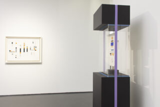 Minimalistische Kunstgalerie mit einem gerahmten abstrakten geometrischen Kunstwerk an der Wand und einer schwarzen Vitrine mit einem kleinen abstrakten Stück.