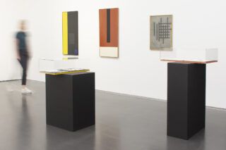 Eine Person geht an zwei Vitrinen auf schwarzen Sockeln in einer Galerie mit abstrakter geometrischer Kunst an weißen Wänden vorbei.