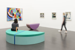 Eine Person sitzt auf einer grünen halbrunden Bank in einer Galerie für moderne Kunst, während eine andere Person an farbenfrohen abstrakten Gemälden an weißen Wänden vorbeigeht.