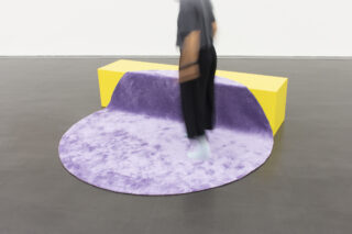 In einer minimalistischen Umgebung steht eine Person auf einem runden lila Teppich, der über einer gelben rechteckigen Bank liegt.