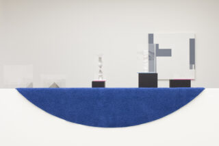 Über einem weißen Sims hängt ein leuchtend blauer halbrunder Teppich, dahinter befinden sich geometrische Skulpturen auf schwarzen Sockeln und an der Wand ein abstraktes Kunstwerk mit grauen Formen.