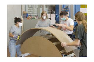 Vier Personen mit Masken und Handschuhen montieren in einer Werkstattumgebung gemeinsam eine gebogene Holzkonstruktion im Innenbereich.