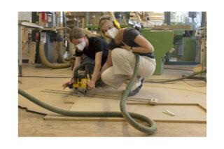 Zwei Personen mit Masken und Gehörschutz schneiden mit einem Elektrowerkzeug Holz auf dem Boden einer Werkstatt, im Hintergrund sind verschiedene Maschinen zu sehen.