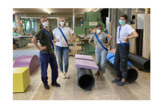 Vier maskierte Personen stehen in einer Werkstatt mit bunten geometrischen Objekten und Maschinen im Hintergrund.