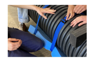 Drei Personen sichern ein großes schwarzes Wellrohr mit einem blauen Gurt und Schaumstoffstützen.