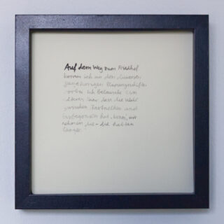 Ein gerahmtes Stück Papier mit handgeschriebenem deutschem Text in schwarzer Tinte, vor einer weißen Wand ausgestellt.