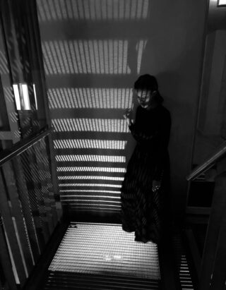 Eine Person in einem langen, dunklen Kleid steht auf einem Gitterboden aus Metall und hält einen Gegenstand, wobei gestreifte Licht- und Schattenmuster auf Wand und Boden geworfen werden.