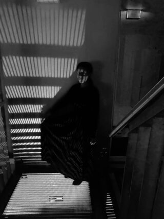 Eine Person in dunkler Kleidung steht auf einer Metalltreppe, bei schwachem Licht werden gestreifte Schatten auf Wand und Boden geworfen.