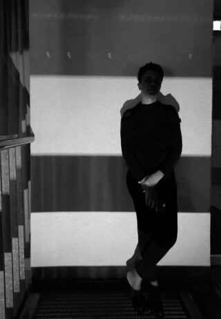 Eine Person steht in einem schwach beleuchteten Bereich vor einer gestreiften Wand und wirft einen Schatten. Die Beleuchtung erzeugt abwechselnd helle und dunkle Bänder in der Szene.
