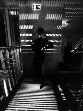 Auf einem Gitterboden mit gestreiften Lichtmustern steht die Silhouette einer Person, darüber hängt an der Wand ein beleuchtetes Notausgangsschild.
