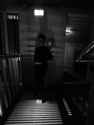 Eine Person steht in einem schwach beleuchteten Bereich auf einem Metallgitterboden. Auf die Wand hinter ihr werden gemusterte Schatten geworfen, und darüber ist ein beleuchtetes Notausgangsschild zu sehen.