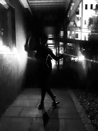 Silhouette einer Person, die nachts mit erhobenem Arm in einem schwach beleuchteten Außenkorridor steht, im Hintergrund sind Lichter und Gebäudestrukturen sichtbar.