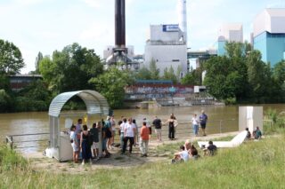 Eine Gruppe von Menschen versammelt sich auf einem Dock am Flussufer in der Nähe einer Industrieanlage. Einige stehen bei einem kleinen überdachten Kiosk, andere sitzen auf dem Rasen.