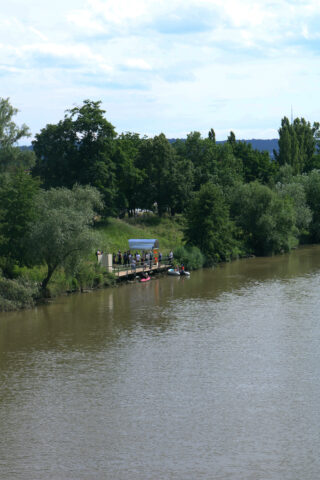Eine Gruppe von Menschen steht auf einem kleinen Steg und bereitet Kajaks oder Kanus am Rande eines braunen Flusses vor, der von grünen Bäumen umgeben ist.