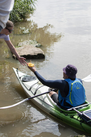 Eine Person in einem Kajak erhält eine Schüssel mit Essen von jemandem, der am Ufer am Wasserrand steht.