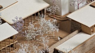 Eine Nahaufnahme eines Architekturmodells, das Gebäude, Wege und Miniaturbäume aus Pappe und anderen Materialien zeigt.