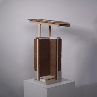 Ein Modell eines Gebäudeabschnitts zeigt eine Stampflehmwand mit einer geschichteten Dachkonstruktion, die auf einem weißen Sockel vor einem schlichten Hintergrund dargestellt ist.