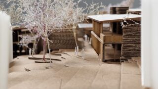 Ein detailliertes Architekturmodell aus Karton zeigt Gebäude, Bäume, Wege und kleine menschliche Figuren in einer Landschaftskulisse.