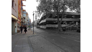 Eine Straßenszene mit mehreren Menschen, die auf dem Bürgersteig neben Geschäften und einem Parkhaus auf der rechten Seite gehen, mit Bäumen und bewölktem Himmel.