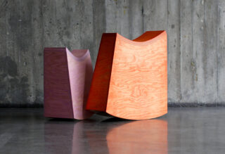 Zwei geometrische Schaukelsitze aus Holz, einer violett und einer orange, stehen auf einem polierten Boden vor einer rauen Betonwand.