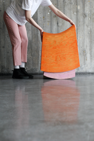 Eine Person in rosafarbener Hose und schwarzen Schuhen hält vor einer Betonwand mit glänzendem Boden ein gebogenes, orangefarbenes Holzobjekt über einem rosafarbenen.