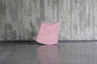 Ein gebogenes Stück hellrosa Sperrholz steht aufrecht vor einer strukturierten Betonwand auf einem polierten grauen Boden.