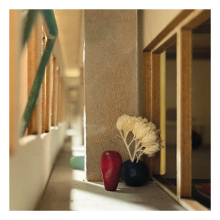 Eine kleine rote Vase und ein runder dunkelblauer Topf mit weißen Trockenblumen stehen in einer Ecke neben einer Wand und großen Fenstern in einem minimal beleuchteten, modern wirkenden Raum.