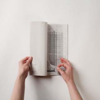 Zwei Hände blättern in einem Buch und bringen eine Seite mit einer Architekturzeichnung eines Gebäudegrundrisses auf weißem Hintergrund zum Vorschein.