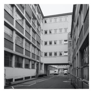 Ein Schwarzweißfoto einer schmalen Gasse zwischen modernen, mehrstöckigen Bürogebäuden, in deren Nähe zwei weiße Lieferwagen vor einem überdachten Durchgang geparkt sind.