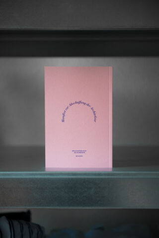 Ein rosa Heft mit blauem Text auf dem Cover steht aufrecht auf einem grauen Regal. Der Text ist bogenförmig angeordnet.