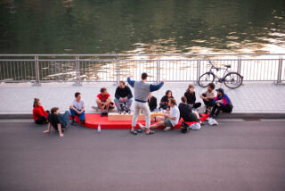 Eine Gruppe von Menschen sitzt im Kreis auf einer roten Bank am Wasser. In der Nähe stehen Snacks bereit und hinter ihnen steht ein Fahrrad am Geländer.