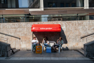Unter einer roten Markise mit der Aufschrift „kaltes Getränk“ sitzen drei Personen auf Plastikstühlen neben einer Steinmauer, vor ihnen stehen Getränke und Kühlboxen.