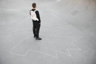 Eine Person in schwarzer Kleidung und einer weißen Weste steht in einem Skatepark aus Beton und blickt weg. Sie steht in der Nähe einer großen abstrakten Form, die mit weißer Umrandung auf dem Boden markiert ist.