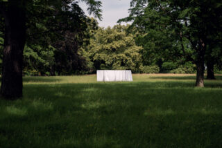 Eine weiße rechteckige Struktur oder ein Objekt steht bei Tageslicht inmitten einer Wiese, umgeben von hohen grünen Bäumen.