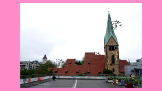 Eine Kirche mit grünem Kirchturm und angrenzendem modernen Gebäude mit rotem Dach, Vögel fliegen am Himmel und im Vordergrund ein Dachgarten, eingerahmt von einem rosa Rand.