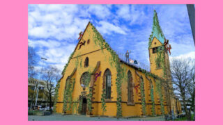 Eine gelbe Kirche mit grünem Efeuschmuck und farbenfrohen Skulpturen von Menschen, die die Wände hochklettern, vor einem bewölkten Himmel mit einem rosa Rand um das Bild.