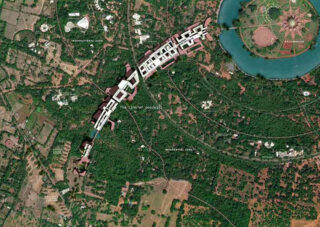 Luftaufnahme eines großen, von Grün umgebenen Gebäudekomplexes, in dessen oberer rechter Ecke ein kreisförmiges Gewässer und Straßen zu sehen sind.