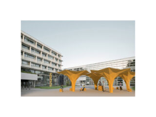 Ein moderner Innenhof mit gelben geometrischen Baldachinstrukturen, spazierenden Menschen und spielenden Kindern; im Hintergrund mehrstöckige Gebäude aus Glas und Beton.