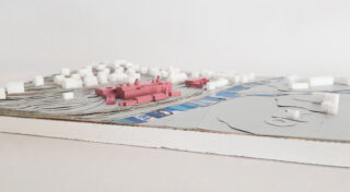 Ein physisches Architekturmodell zeigt geschichtetes Gelände, weiße Blockgebäude, blaue Straßen und rosafarbene Strukturen in der Mitte.