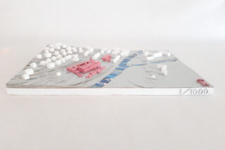 Ein maßstabsgetreues Architekturmodell mit weißen und rosafarbenen Gebäuden, einem blauen Fluss und Höhenlinien, dargestellt auf einer rechteckigen Basis mit der Bezeichnung 1/1000.