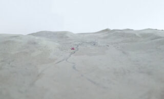 Eine Nahaufnahme eines blassen, strukturierten Landschaftsmodells mit subtilen Höhenunterschieden und einem kleinen rosa Objekt in der Nähe der Mitte.