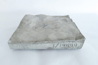Ein rechteckiges Betonmodell mit einer erhabenen topografischen Landschaft, auf dessen Seite „1/10000“ eingeprägt ist, fotografiert vor einem weißen Hintergrund.