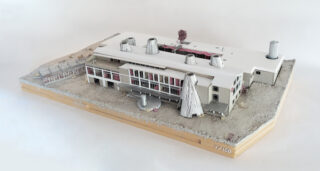 Ein detailliertes Architekturmodell eines modernen Gebäudekomplexes mit zylindrischen Dachkonstruktionen auf einem rechteckigen Holzsockel, gekennzeichnet mit einem Maßstab von 1/100.